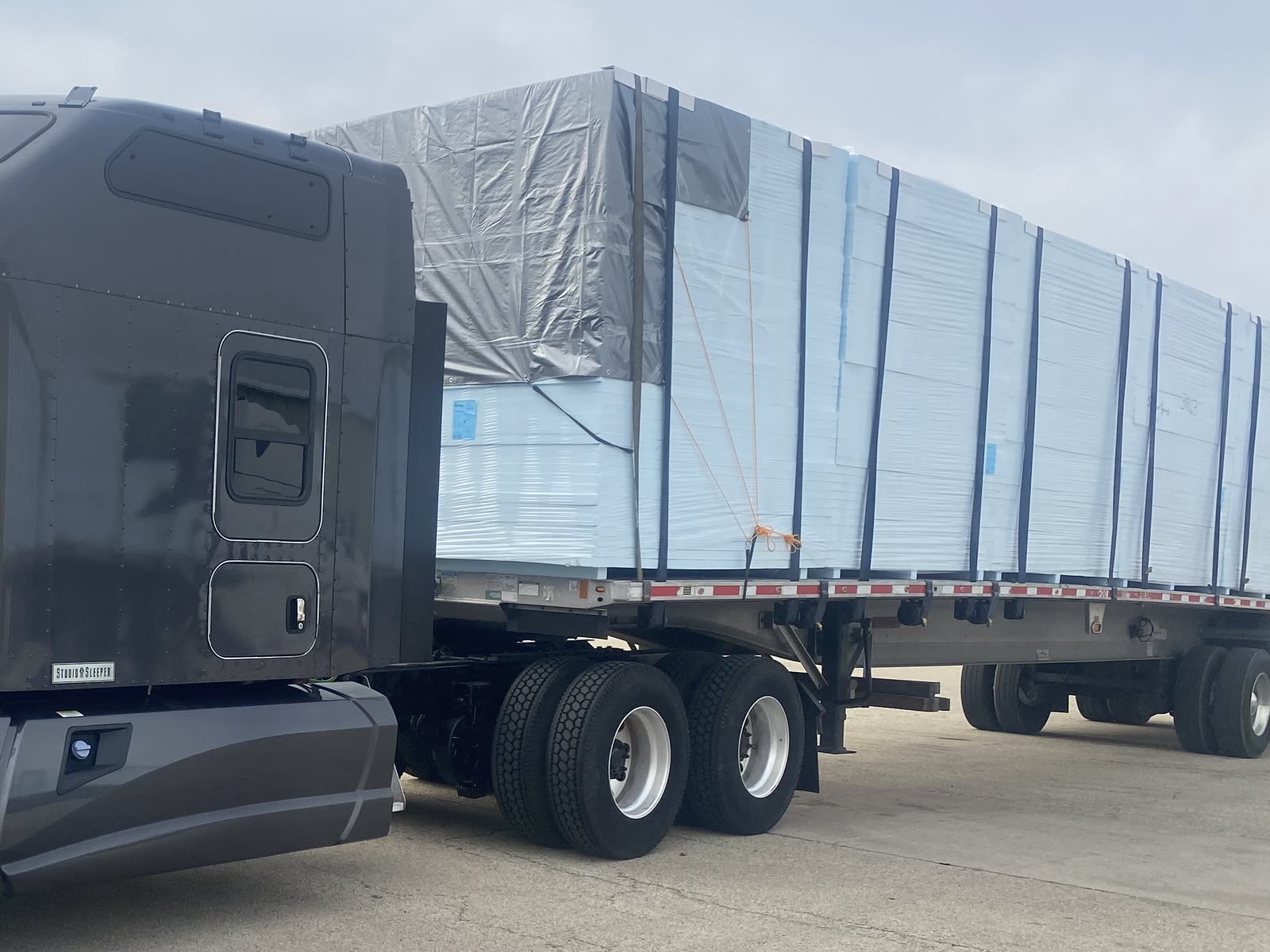 Flatbed trailer stacked with wrapped and strapped building-material panels behind a dark gray tractor