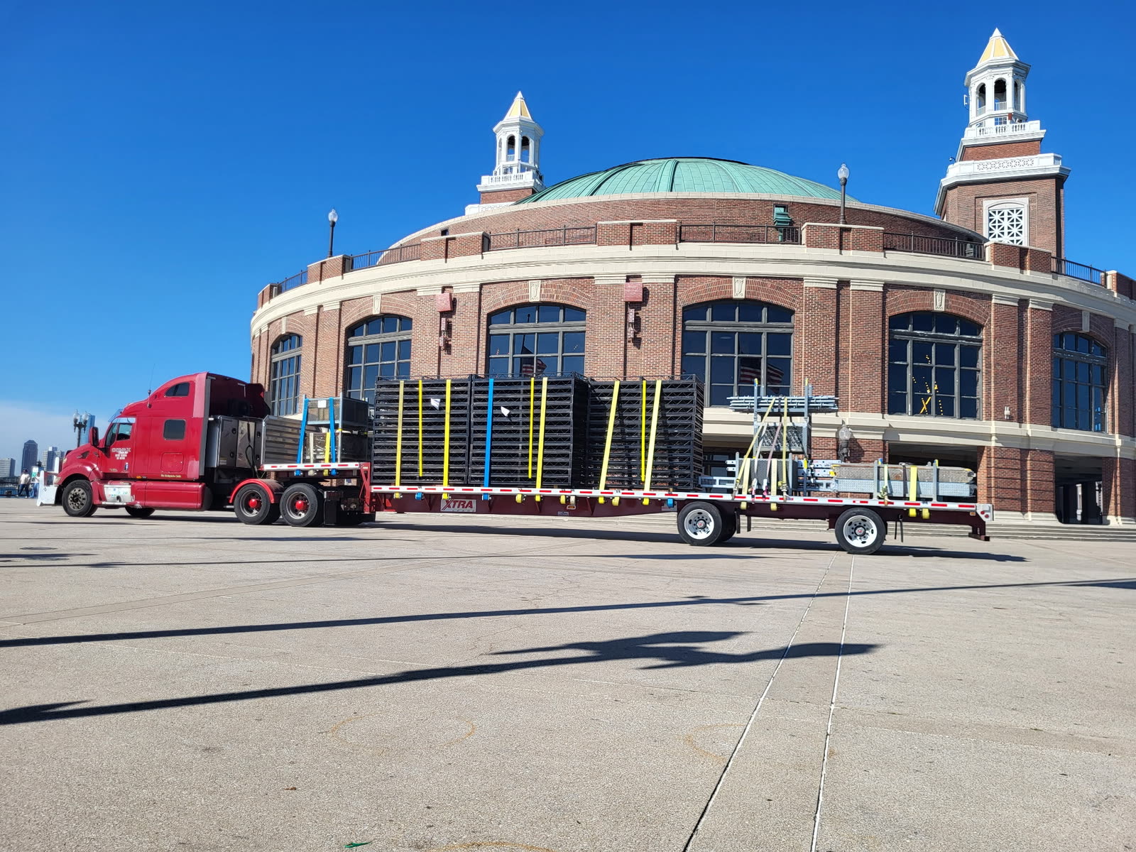Red tractor pulling a flatbed loaded with strapped event staging equipment in front of a domed brick building