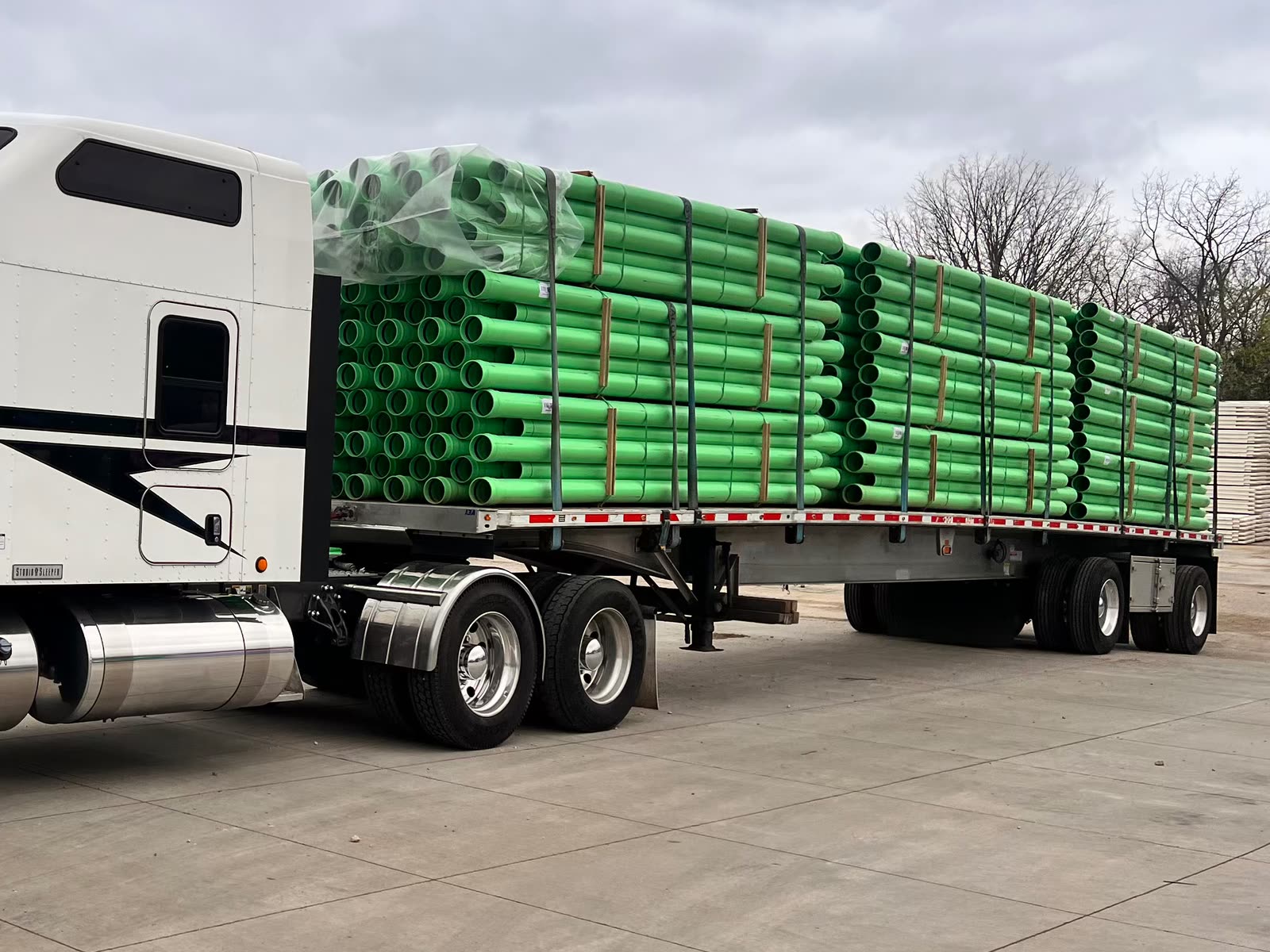 Flatbed trailer loaded with bundles of green plastic pipe behind a white tractor