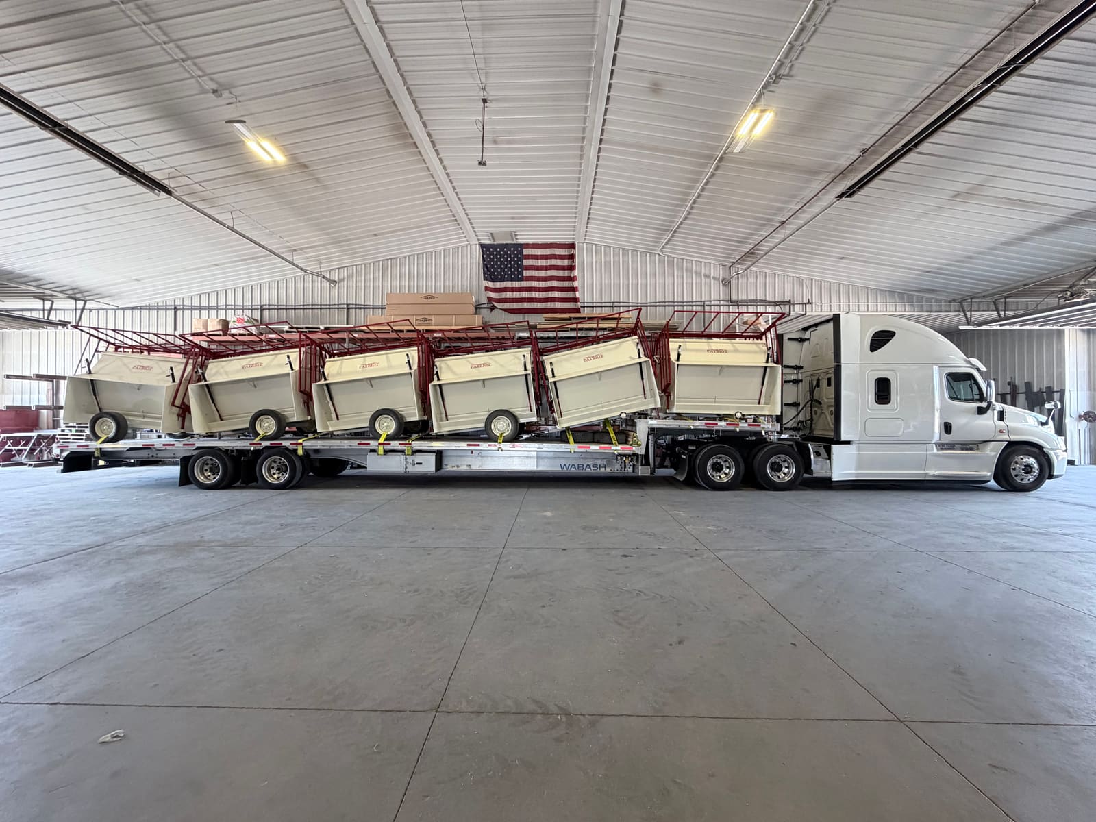 White Covax truck with a loaded step-deck trailer parked inside a warehouse, an American flag hanging on the wall behind it